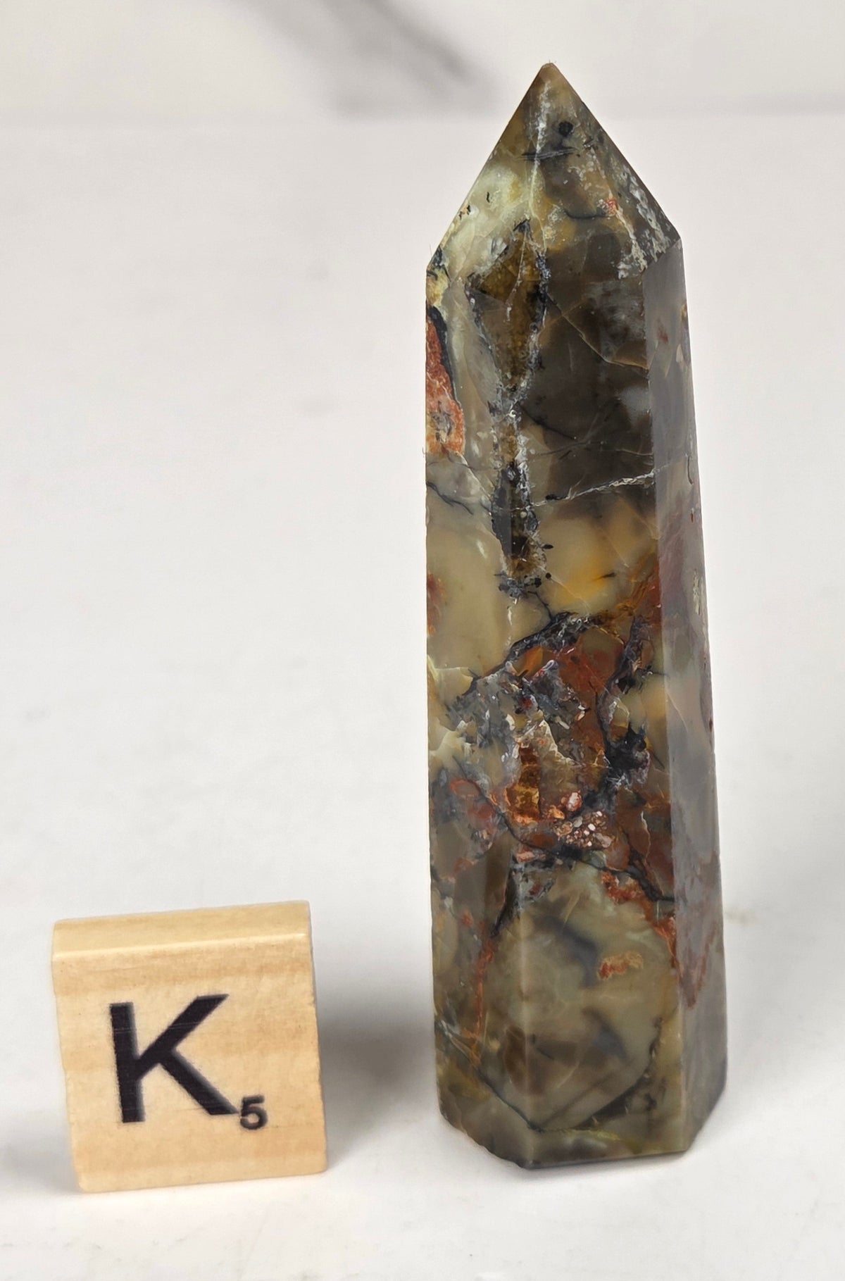 Volcano Agate polished point with tan and black coloring with a scrabble letter against a marbled background
