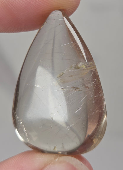 Clear quartz teardrrop shaped crystal with gold rutile inclusions held between fingers