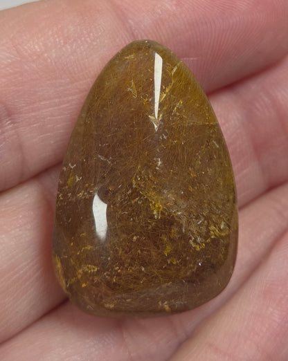 Clear quartz teardrrop shaped crystal with gold rutile inclusions held between fingers