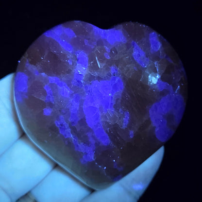 Hand holding a white crystal heart with purple coloring under UV light against a black background