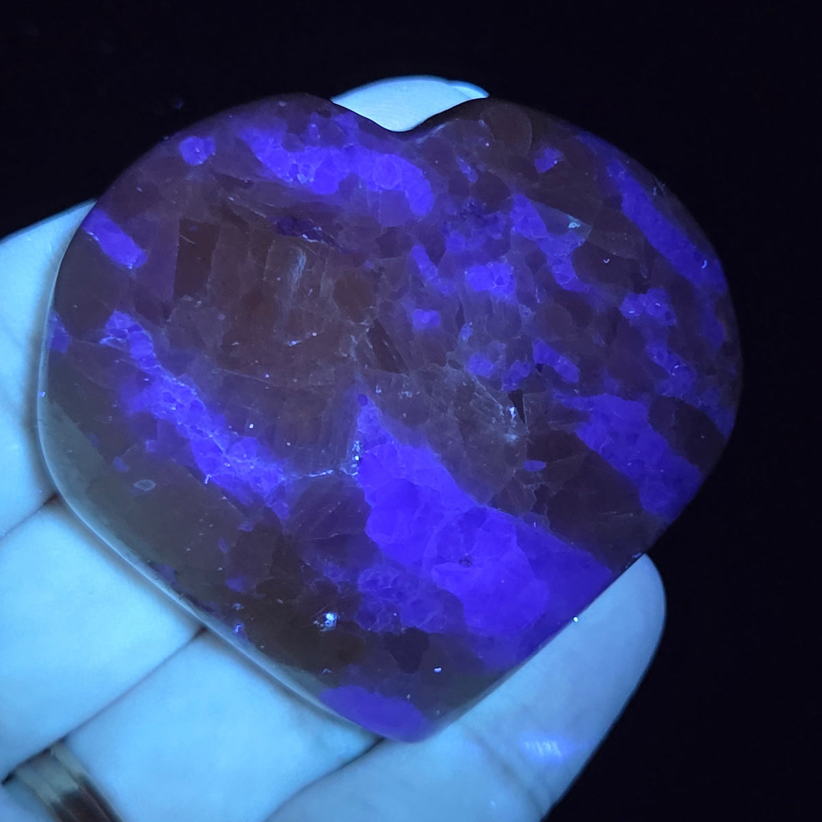 Hand holding a white crystal heart with purple coloring under UV light against a black background