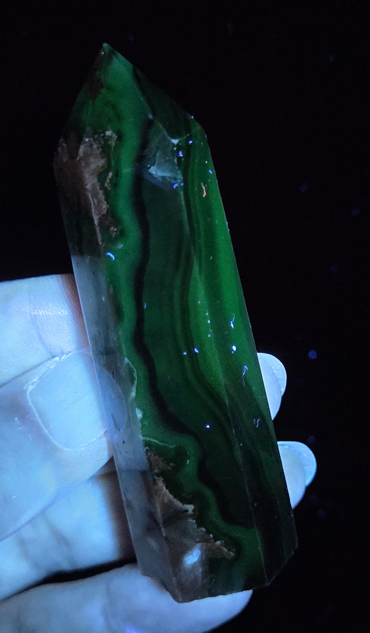 Volcano Agate polished point being held in a hand showing green  color under UV lighting against a  black background