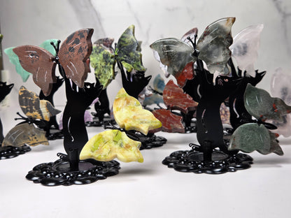 A collection of decorative butterfly wall art pieces displayed on a white surface against a light gray marble-patterned background. Each butterfly features a black silhouette cat body holding a uniquely colored stone wing, including shades of brown, green, yellow, red, and gray.