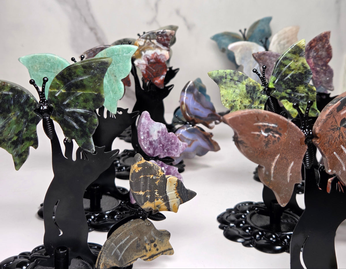 A collection of decorative butterfly wall art pieces mounted on black metal stands, featuring various colored stone wings in green, brown, purple, blue, and yellow, displayed against a light marble-patterned background.