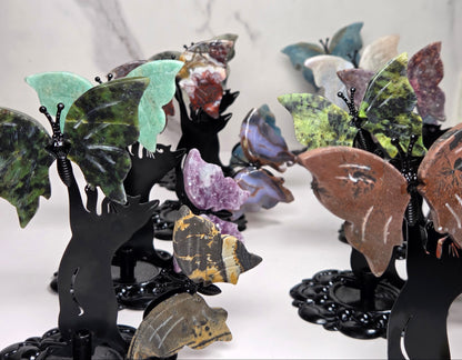 A collection of decorative butterfly wall art pieces mounted on black metal stands, featuring various colored stone wings in green, brown, purple, blue, and yellow, displayed against a light marble-patterned background.