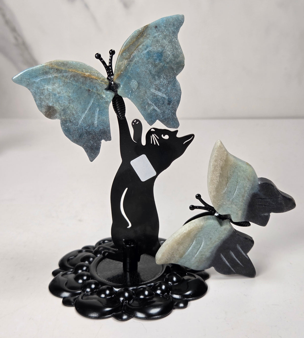 A decorative black metal figurine of a cat reaching up to a large blue butterfly, with a smaller matching butterfly beside it, displayed on a light surface against a marble-patterned background.