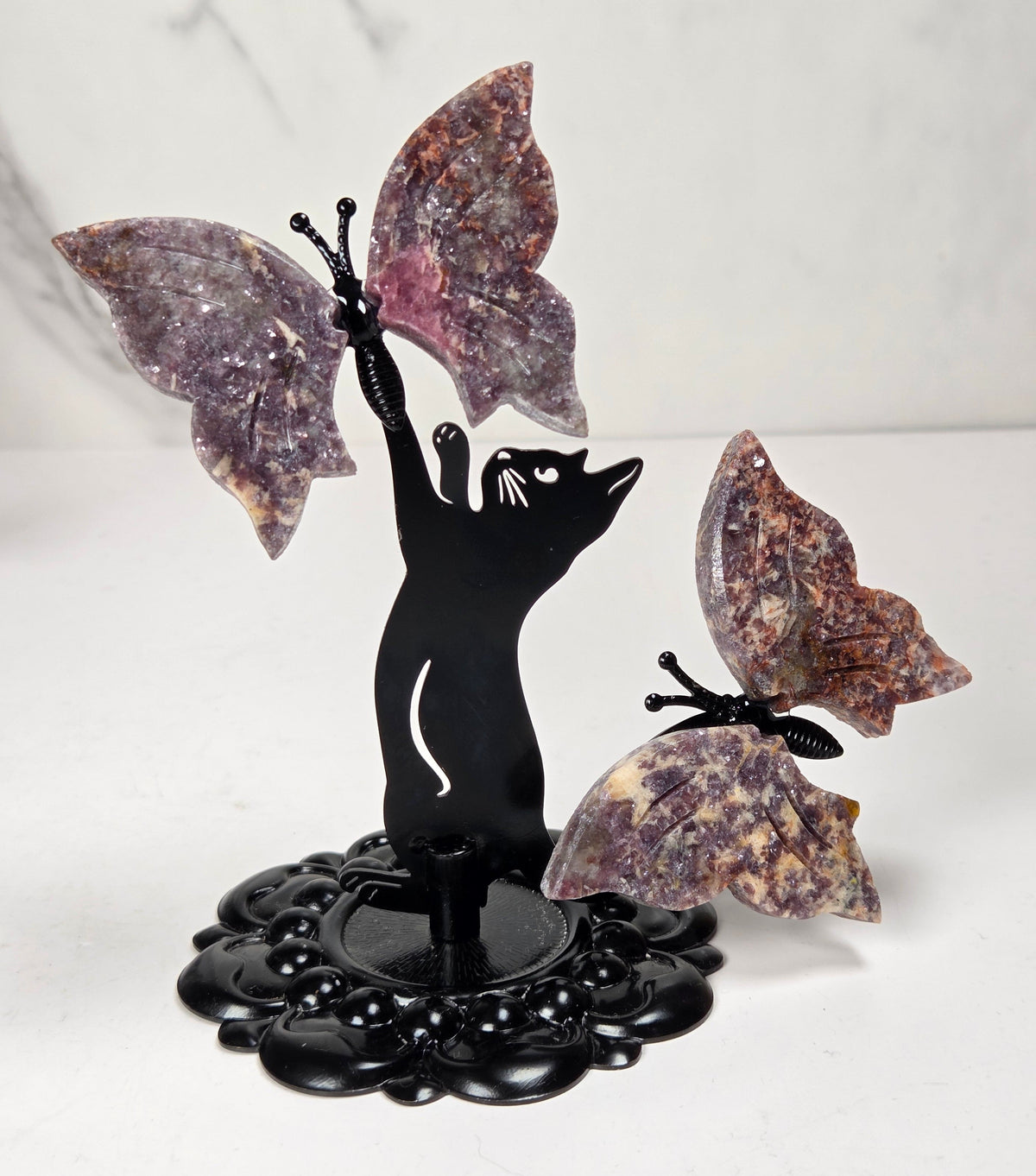 A decorative black metal sculpture of a cat reaching upward with one winged butterfly attached, alongside another butterfly on the right, all mounted on a black floral base against a light marble-patterned background.