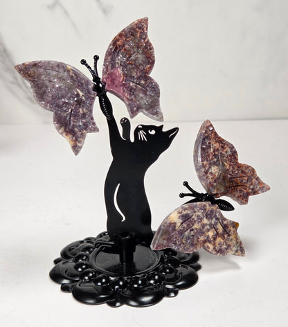 A decorative black metal sculpture of a cat reaching upward with one winged butterfly attached, alongside another butterfly on the right, all mounted on a black floral base against a light marble-patterned background.