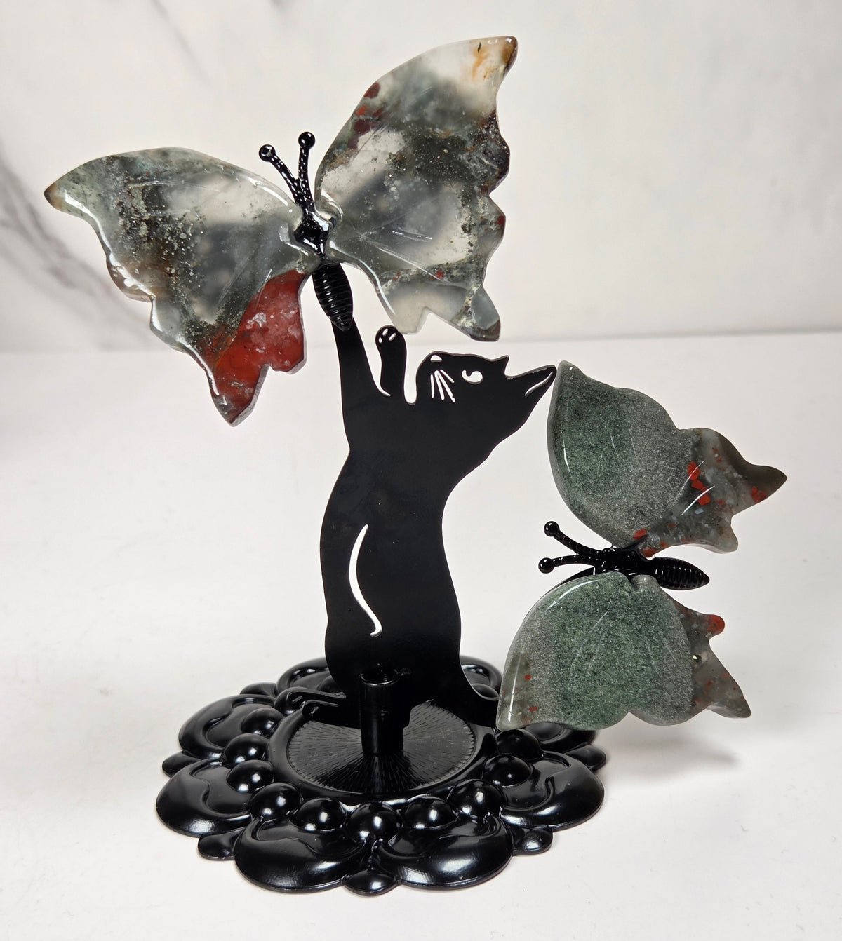 A decorative sculpture featuring a black cat silhouette reaching upward to touch two butterfly wings made from polished stone. One butterfly is gray with white marbling and a red patch, and the other is green with small red inclusions.