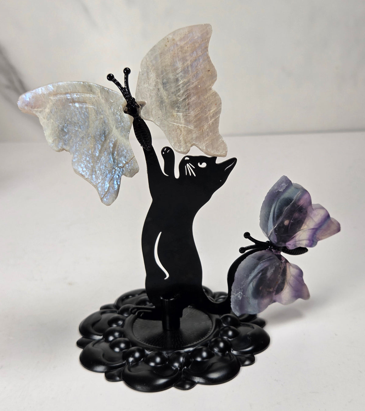 A decorative black metal figurine shaped like a cat reaching upward with one wing, while a second purple-toned butterfly rests beside it. The butterflies appear to be made of iridescent stone or resin, and the base is a dark ornate pedestal.