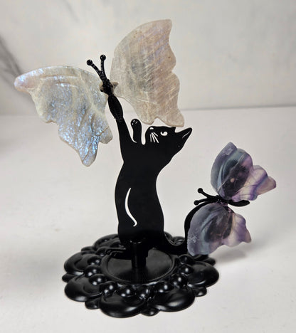 A decorative black metal figurine shaped like a cat reaching upward with one wing, while a second purple-toned butterfly rests beside it. The butterflies appear to be made of iridescent stone or resin, and the base is a dark ornate pedestal.