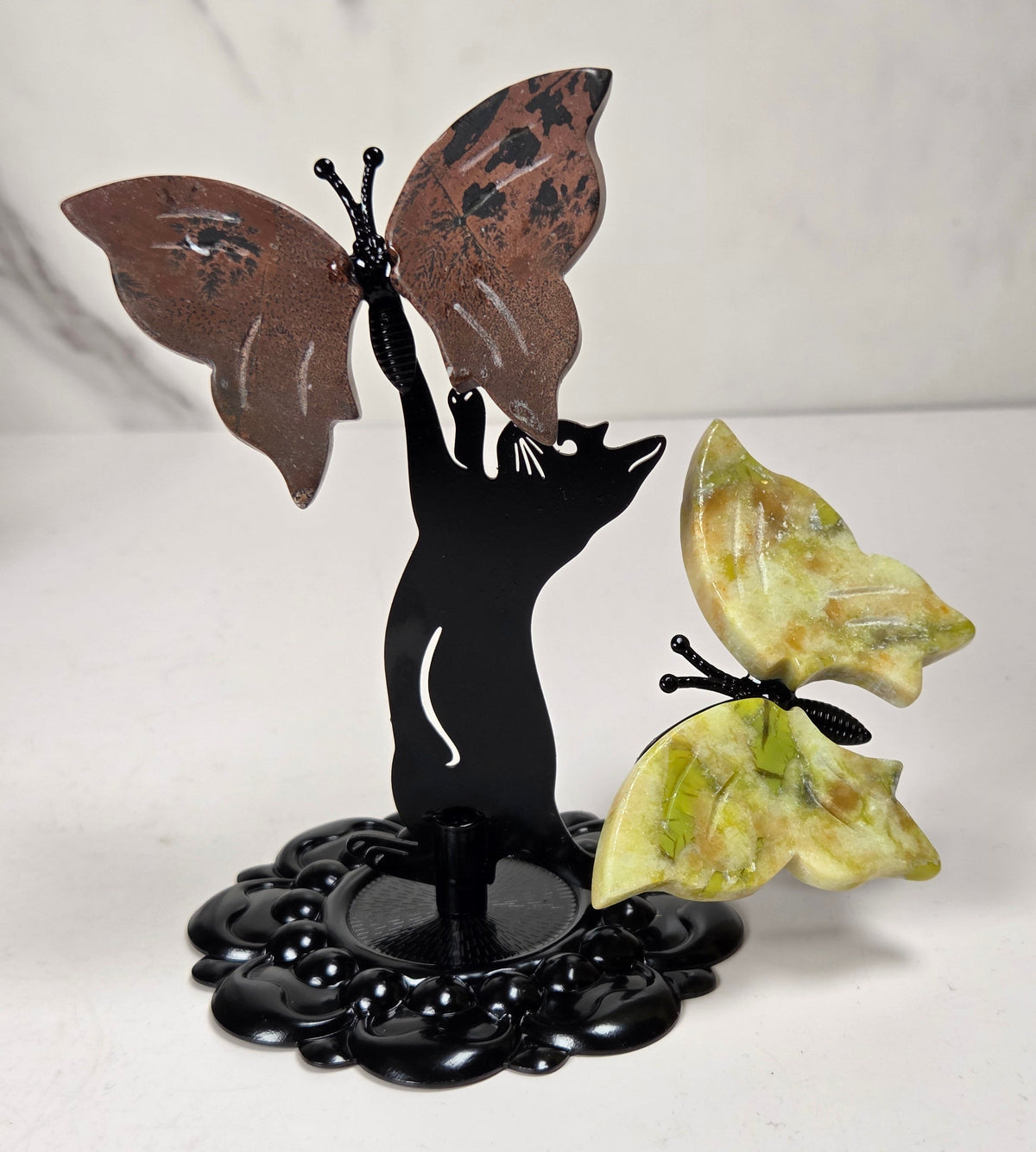 A decorative metal and stone butterfly sculpture featuring a black cat silhouette holding a brown butterfly, with a green marbled butterfly perched beside it on an ornate black base.