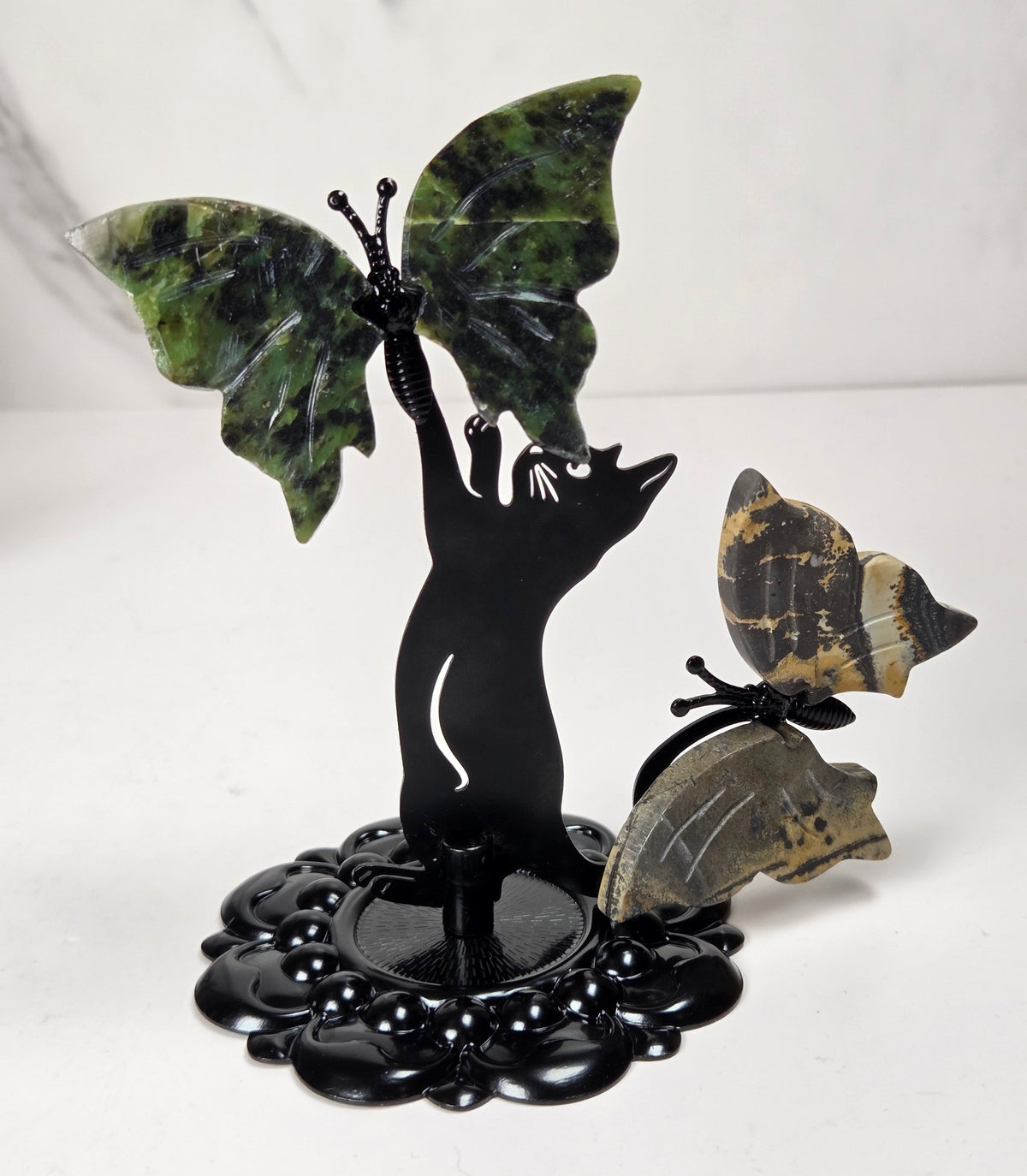 A decorative black metal sculpture of a cat with butterfly wings, featuring green marbled stone butterfly wings and two smaller patterned butterfly wings, displayed on a white surface against a light gray marble background.