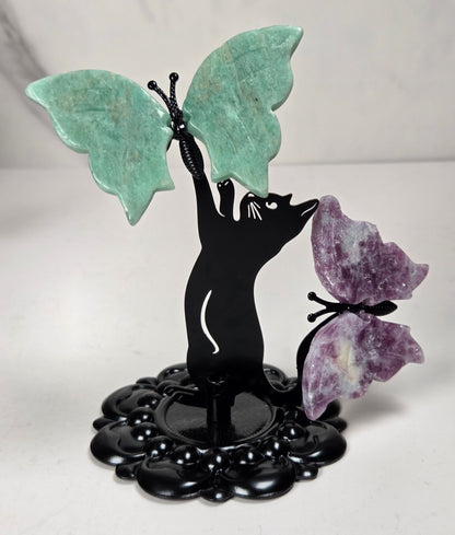 A decorative black metal figurine of a cat perched on a leafy base, with two butterfly wings made from polished stone—one turquoise green and one purple amethyst—attached to the figure, set against a light marble-patterned background.
