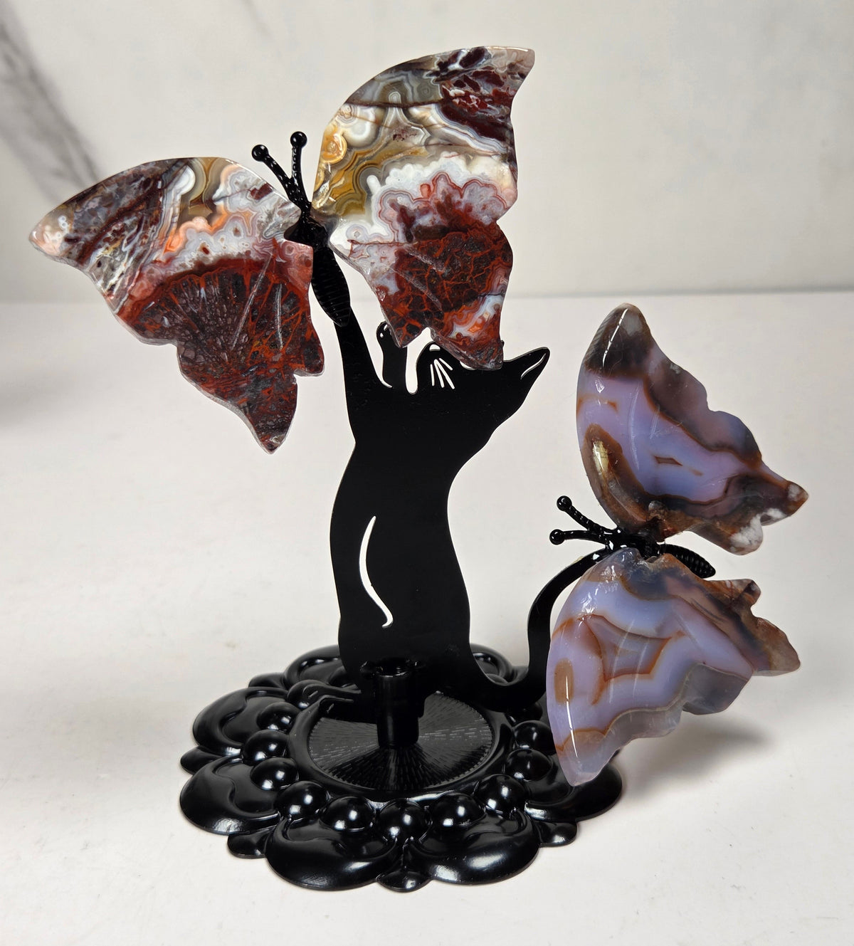 A decorative black metal stand shaped like a cat silhouette, holding three agate butterfly wings. The butterflies feature natural marbled patterns in earthy tones of brown, red, white, and purple, set against a light marble-patterned background.
