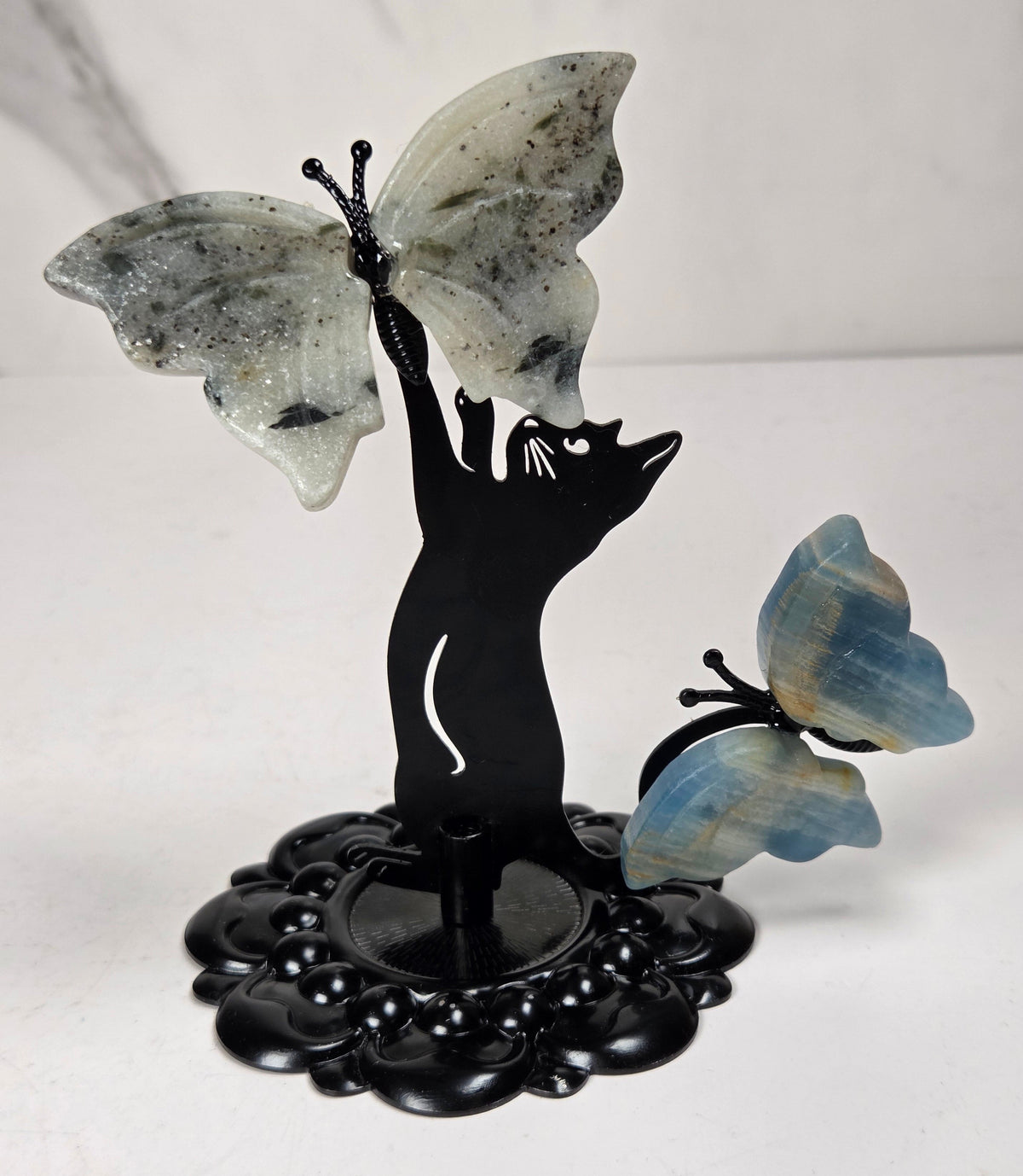 A decorative figurine of a black cat silhouette holding a large pale gray butterfly above its head, with a smaller blue-and-gray butterfly perched on the right, all mounted on an ornate black base against a light neutral background.