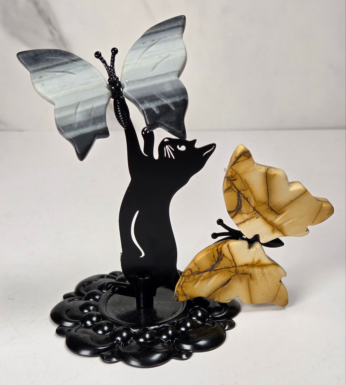 A decorative tabletop sculpture featuring a black silhouette of a cat with wings spread, holding a black-and-white butterfly, alongside a tan-and-gold butterfly mounted on a black base.