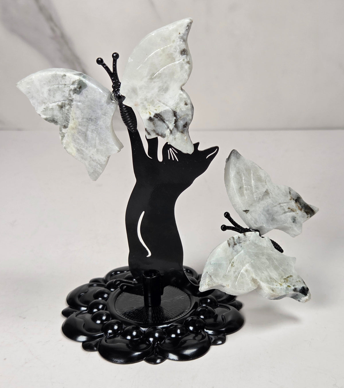 A decorative butterfly sculpture featuring a black cat silhouette holding two large translucent white crystal butterflies, mounted on a black floral base against a light gray marble-patterned background.