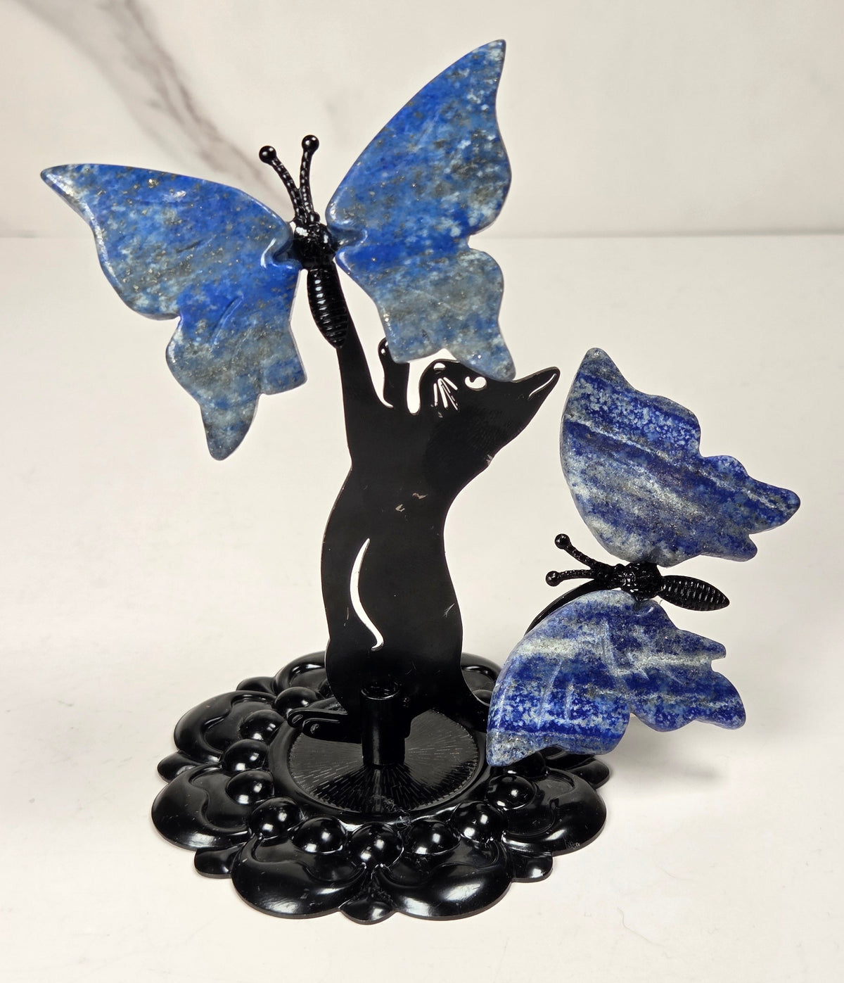 A decorative sculpture of a black cat silhouette with large blue lapis lazuli butterfly wings, mounted on a glossy black floral base against a plain light background.