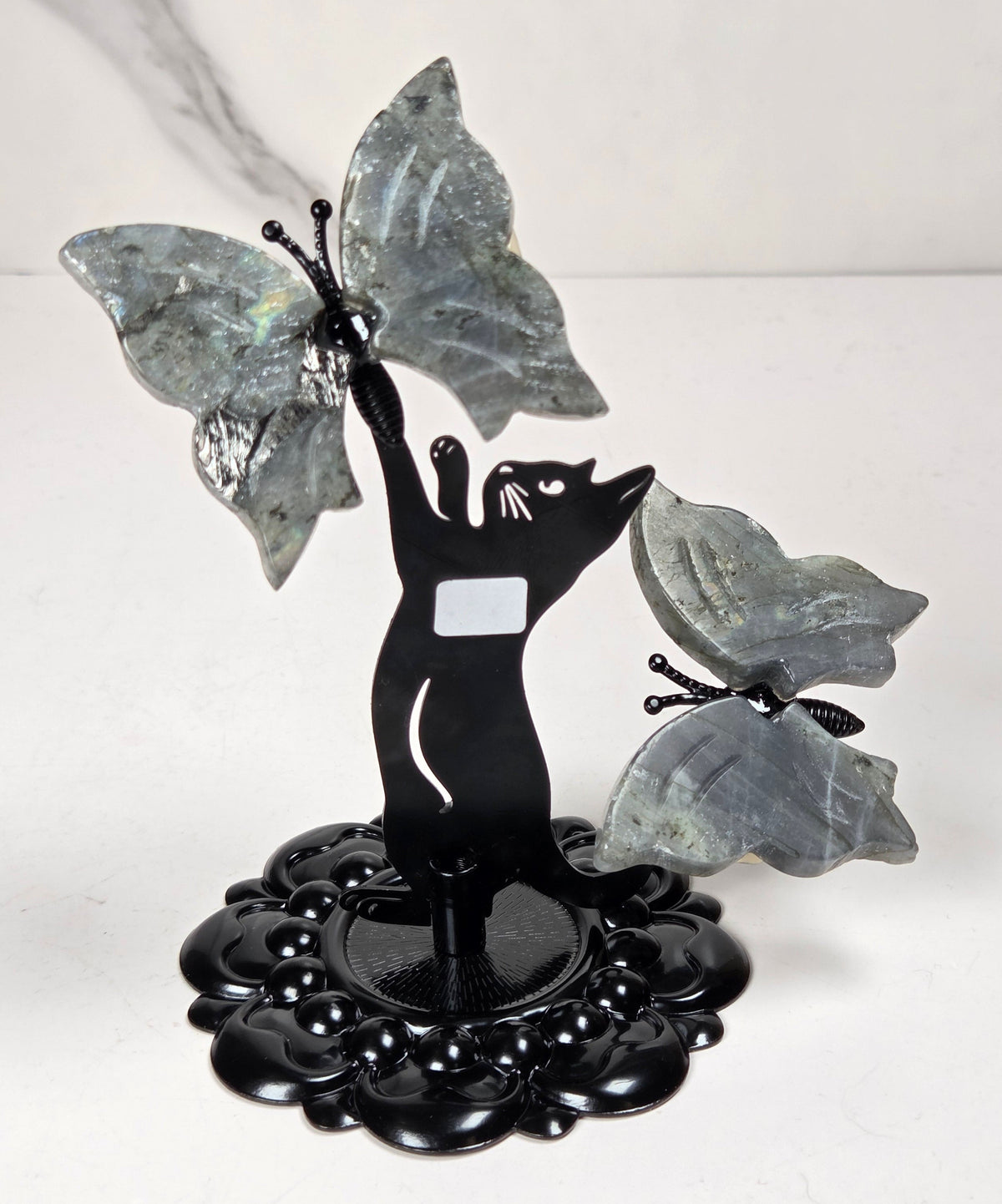 A decorative black metal sculpture of a cat reaching upward toward two large gray stone butterflies, mounted on a round black base against a light marble-patterned background.