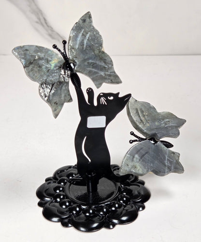 A decorative black metal sculpture of a cat reaching upward toward two large gray stone butterflies, mounted on a round black base against a light marble-patterned background.