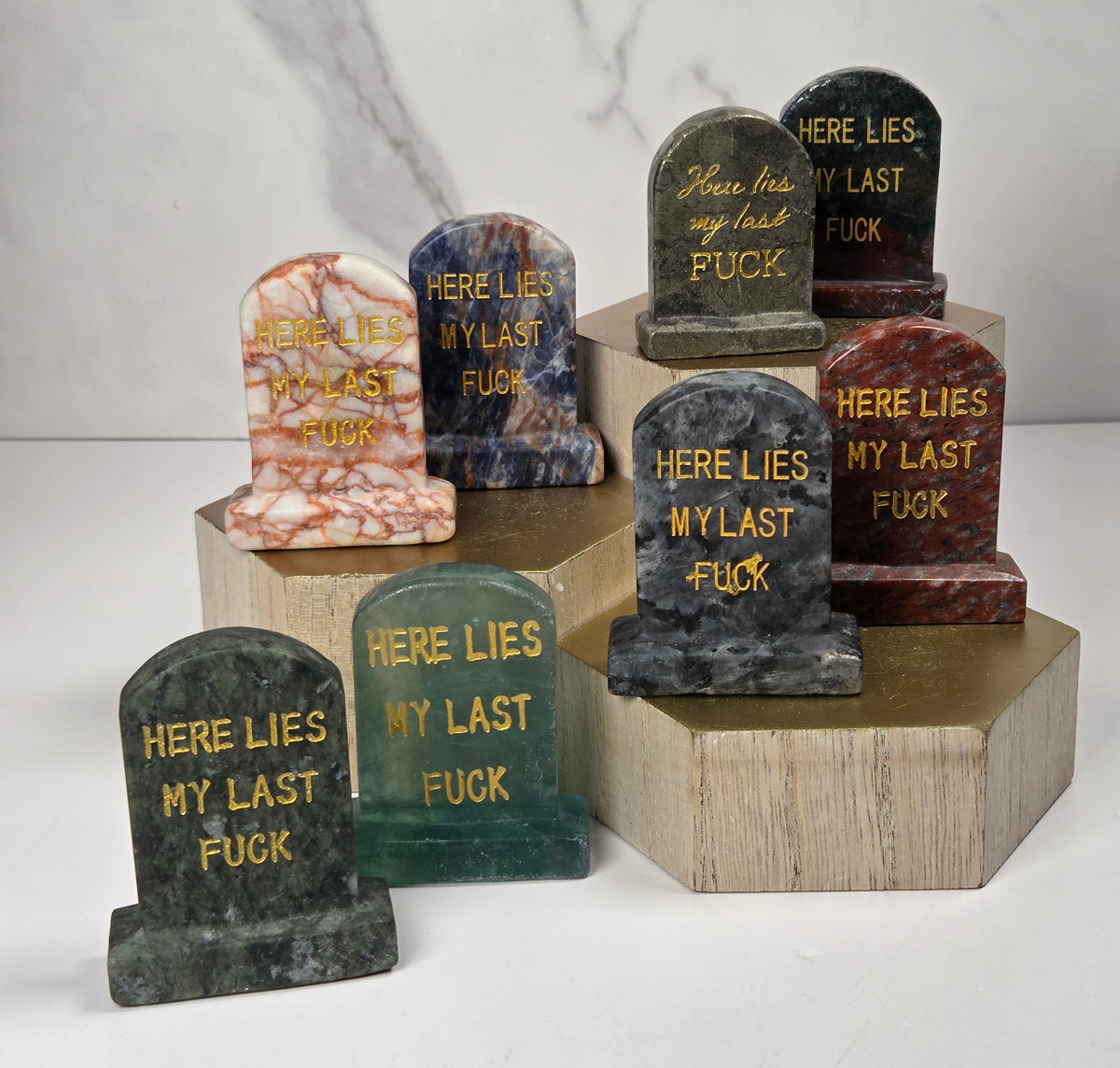 Collection of various colored crystal tombstone carvings with gold lettering on a wooden stand against a white background