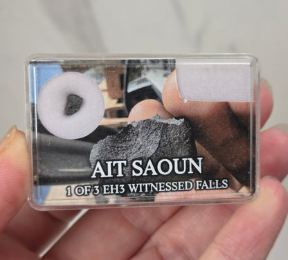hand holding a clear box with a meteorite specimen against a white background