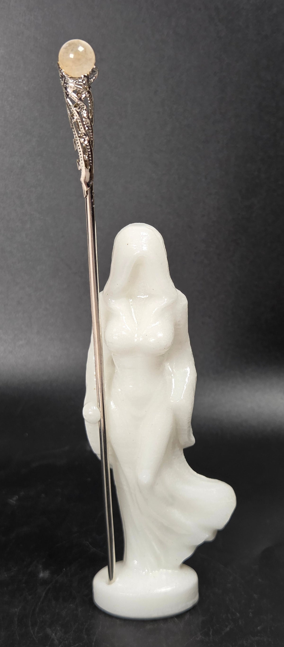 White crystal carving of a faceless woman wearing a robe with a metal wand against a black background