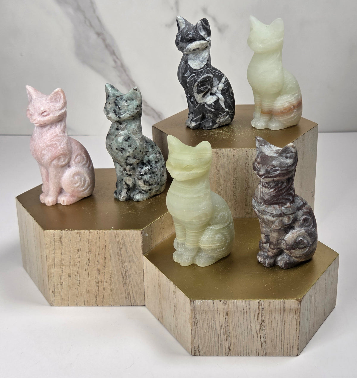 Collection of crystal carvings shaped like a cat, in a variety of colors and materials, all sitting on a wooden shelf against a marbled background