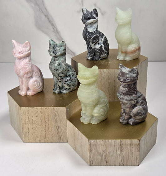 Collection of crystal carvings shaped like a cat, in a variety of colors and materials, all sitting on a wooden shelf against a marbled background