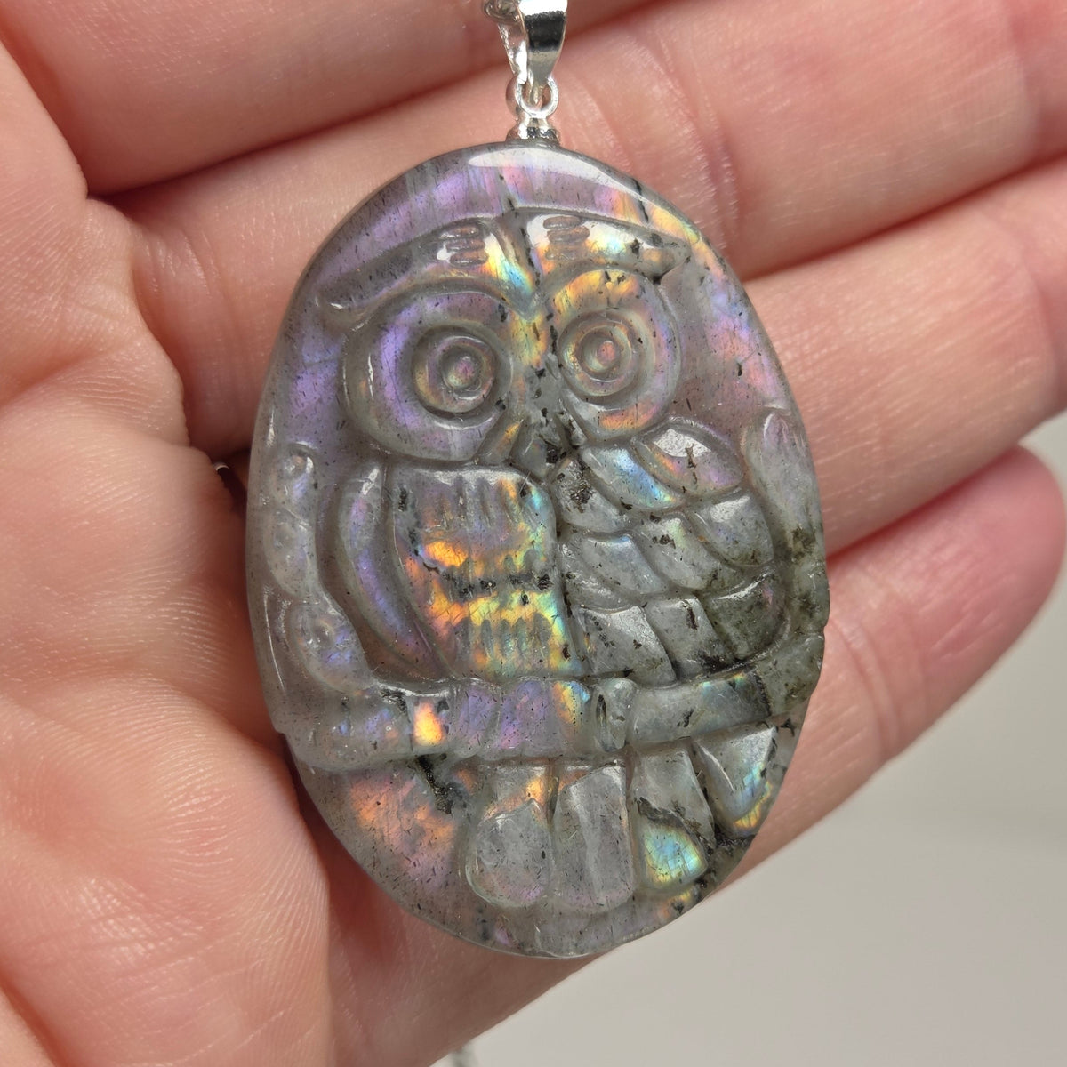 Hand holding a grey crystal owl shaped pendant with rainbow flash on a silver chain against a white background