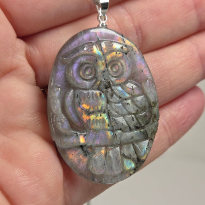 Hand holding a grey crystal owl shaped pendant with rainbow flash on a silver chain against a white background