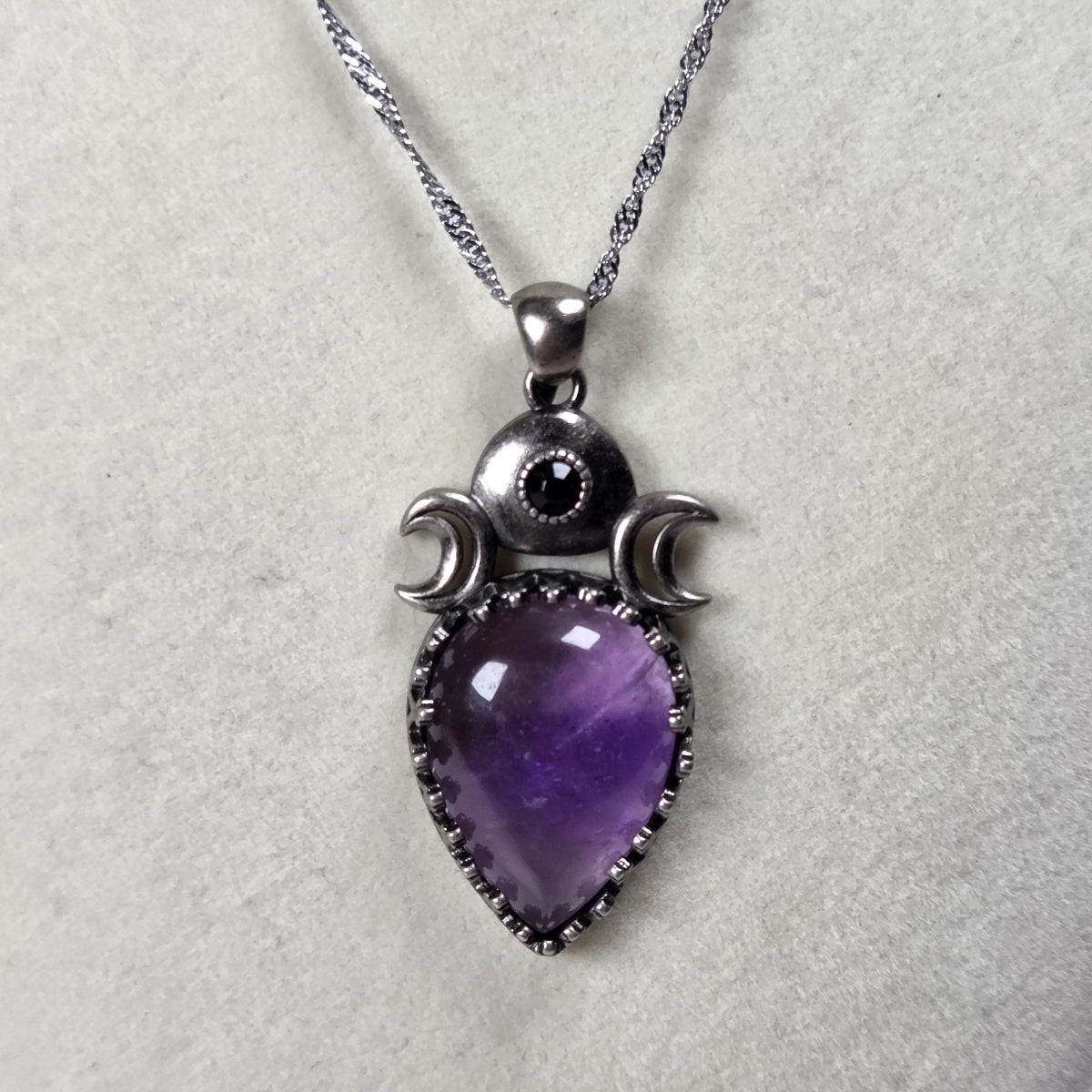 A silver pendant withe a purple crystal cabachon with silver moon accents, on a silver chain against a white background