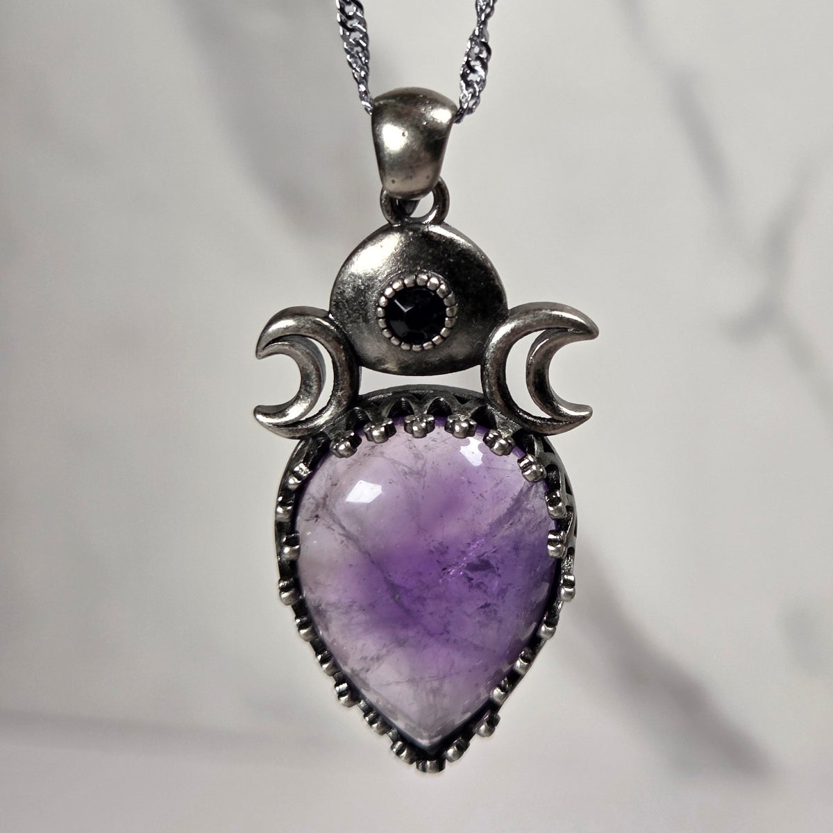A silver pendant withe a purple crystal cabachon with silver moon accents, on a silver chain against a white background