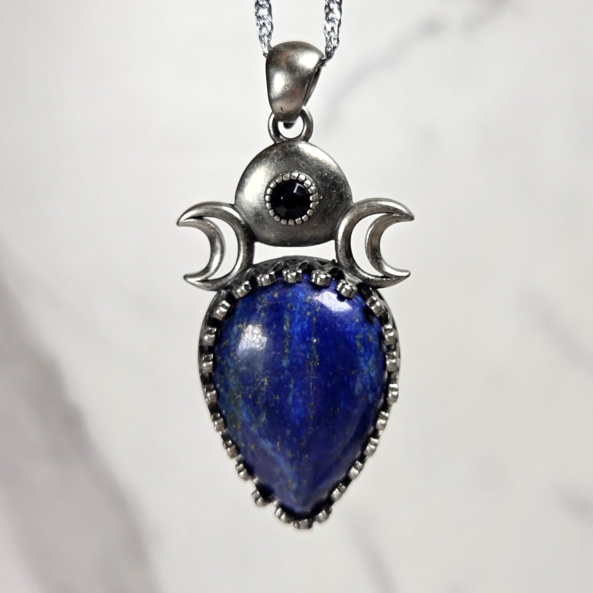 A silver pendant withe a blue crystal cabachon with silver moon accents, on a silver chain against a white background
