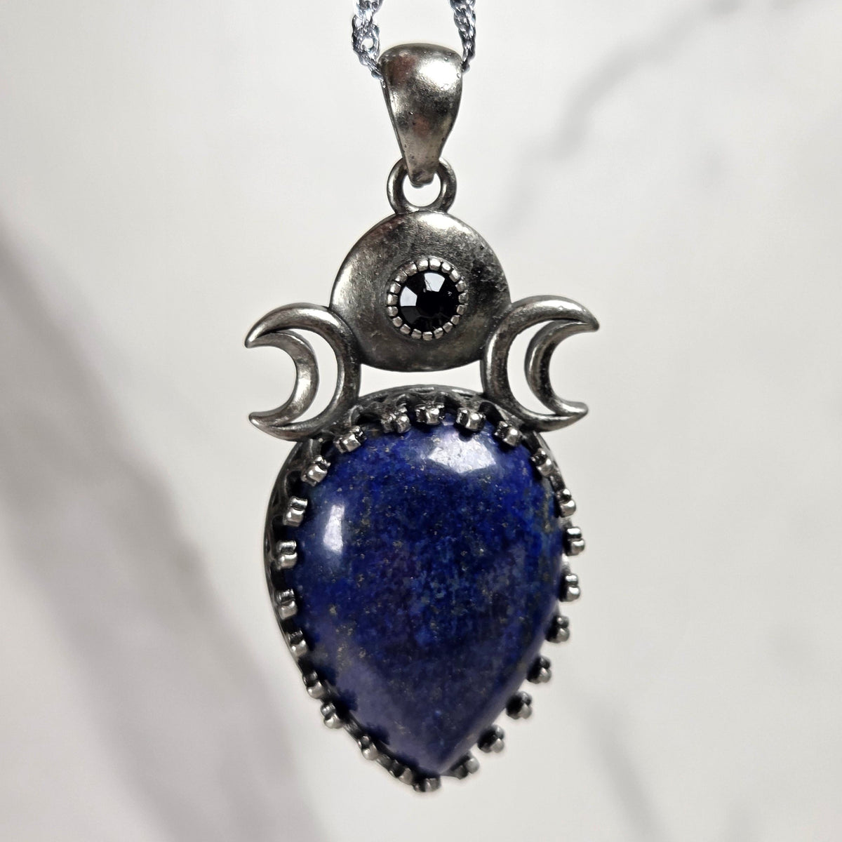 A silver pendant withe a blue crystal cabachon with silver moon accents, on a silver chain against a white background