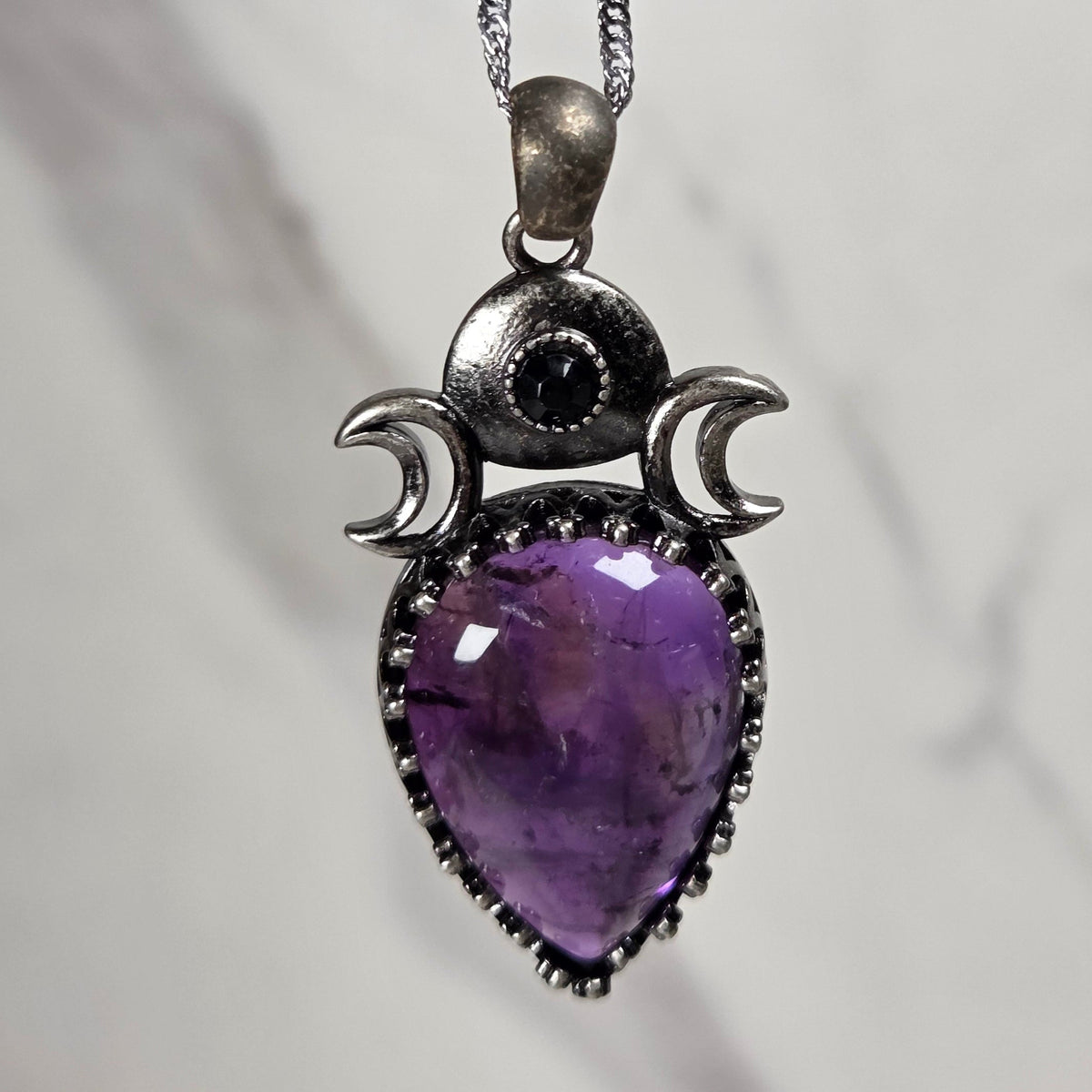 A silver pendant withe a purple crystal cabachon with silver moon accents, on a silver chain against a white background