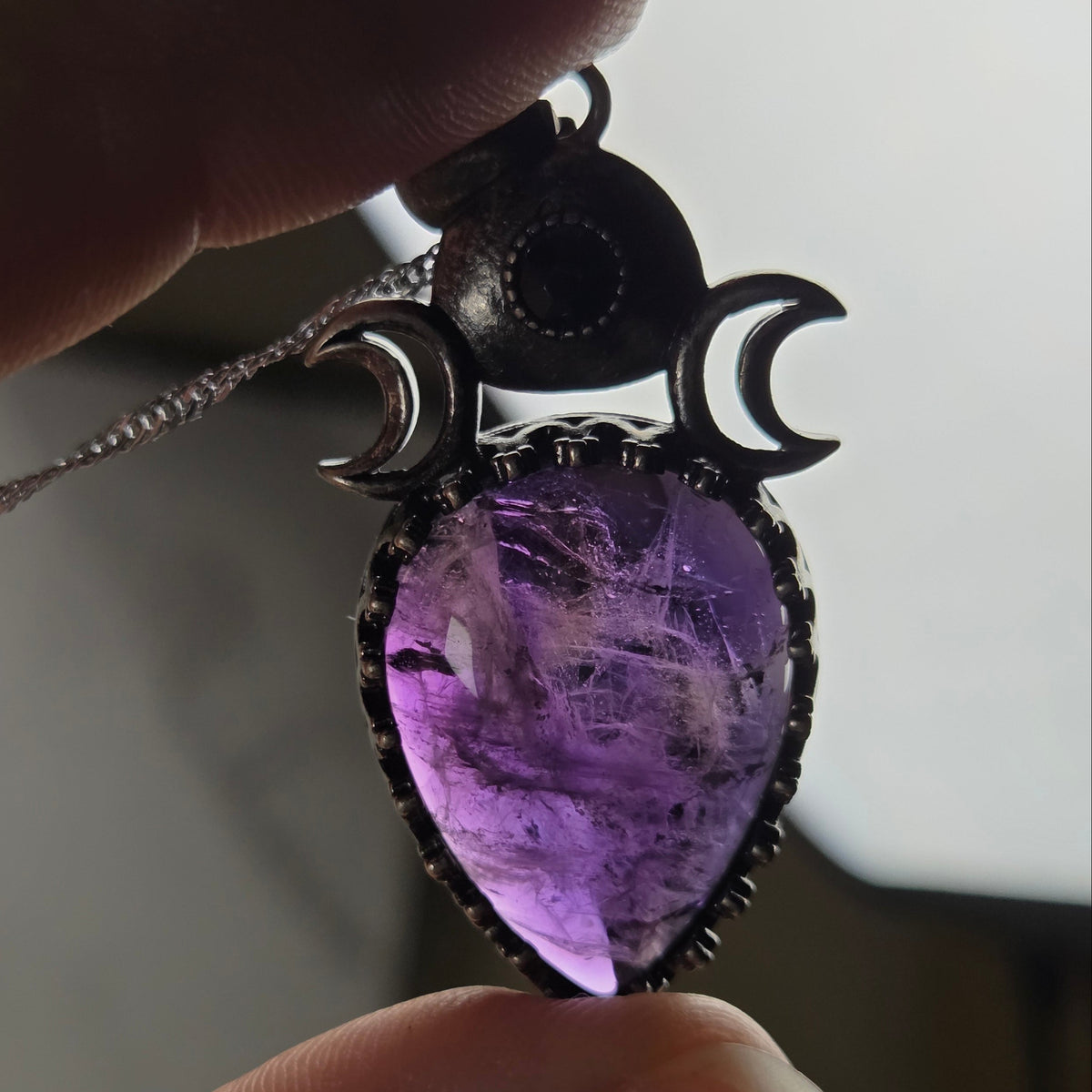 Hand holding a silver pendant withe a purple crystal cabachon with silver moon accents, on a silver chain against a white background