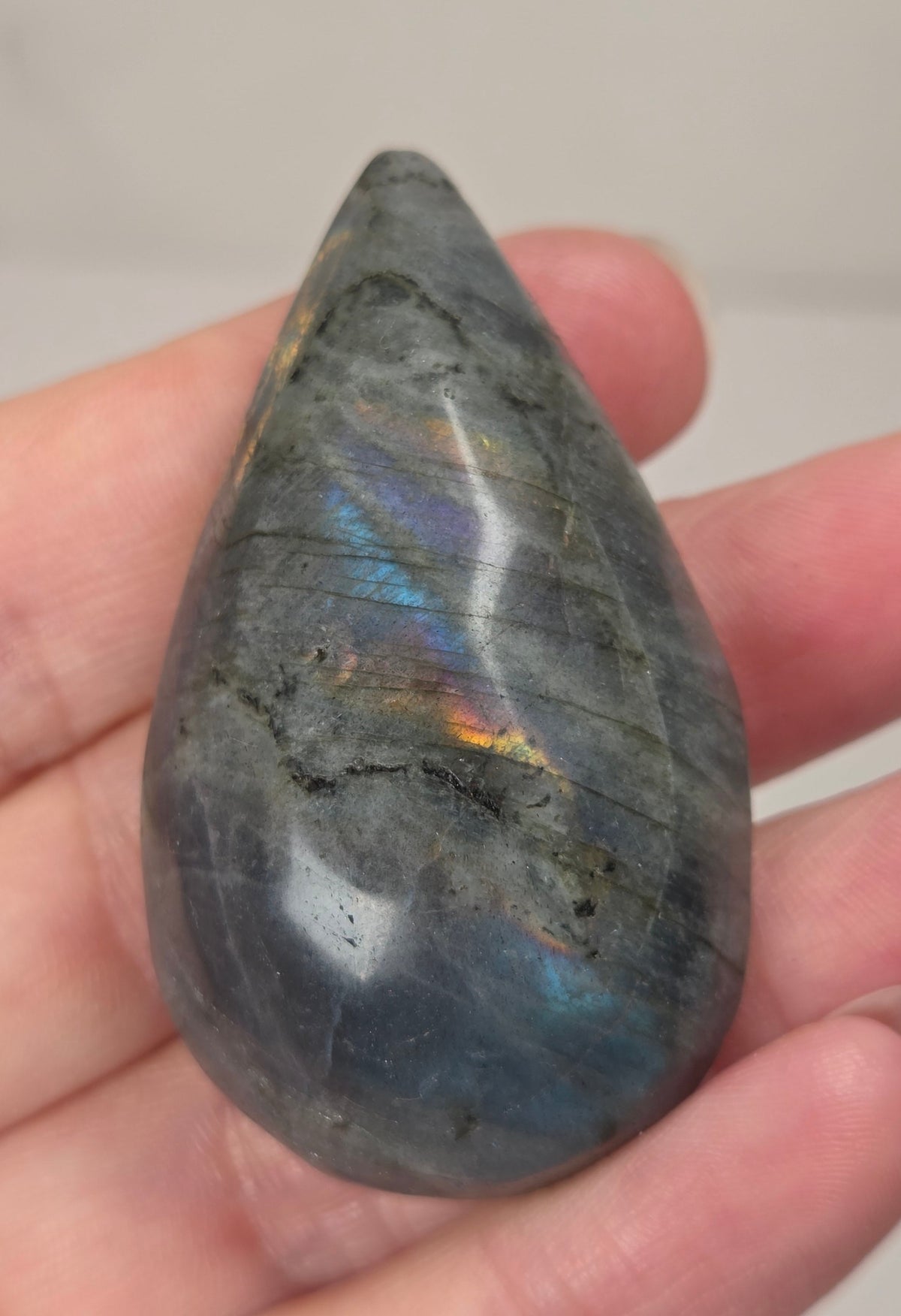 Hand holding a grey labradorite carved raindrop shaped carving with rainbow coloring against a white background