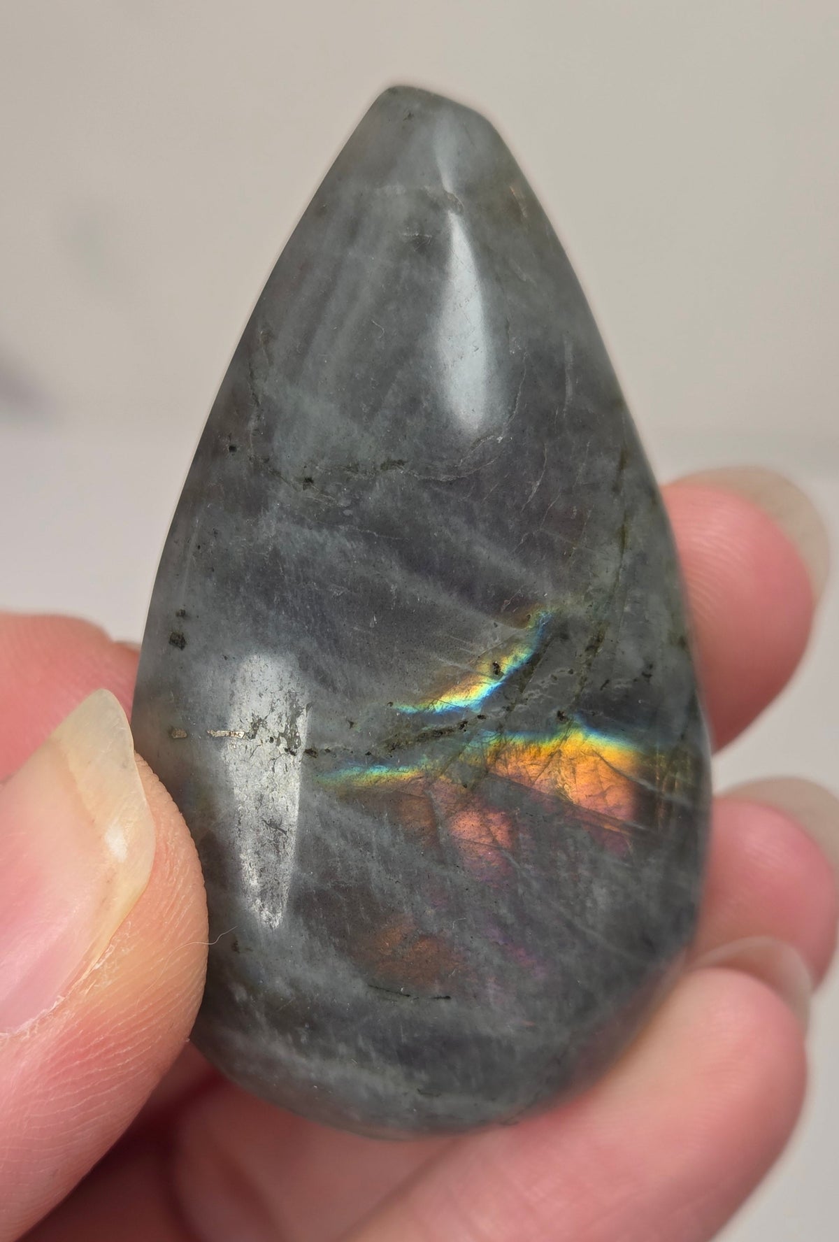 Hand holding a grey labradorite carved raindrop shaped carving with rainbow coloring against a white background