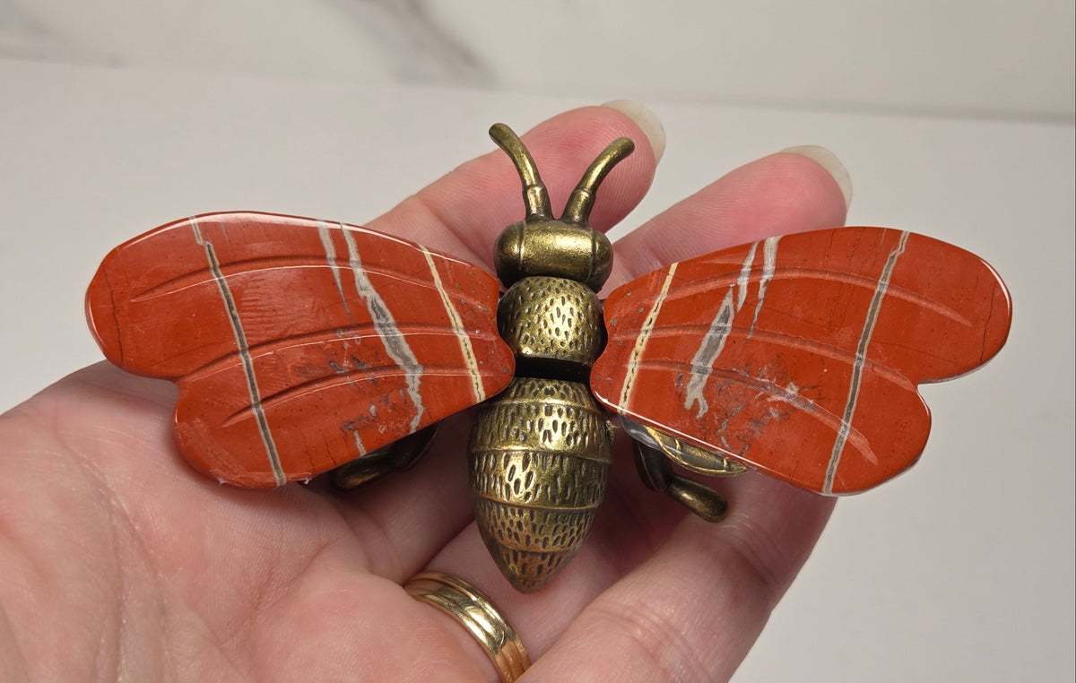A close-up of a hand holding a vintage-style brass bumblebee brooch. The brooch features detailed wing and body textures with a warm gold and bronze metallic finish.