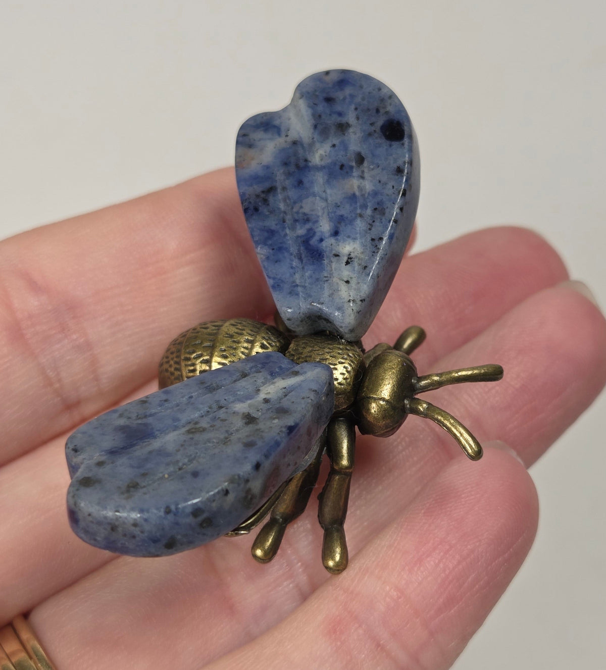 A close-up of a hand holding a vintage-style brass bumblebee brooch. The brooch features detailed wing and body textures with a warm gold and bronze metallic finish.