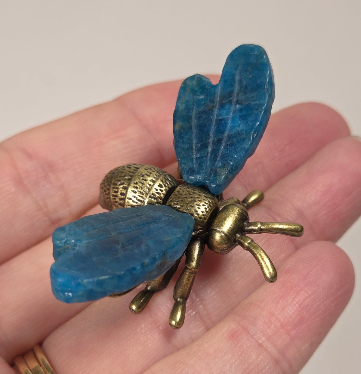 A close-up of a hand holding a vintage-style brass bumblebee brooch. The brooch features detailed wing and body textures with a warm gold and bronze metallic finish.