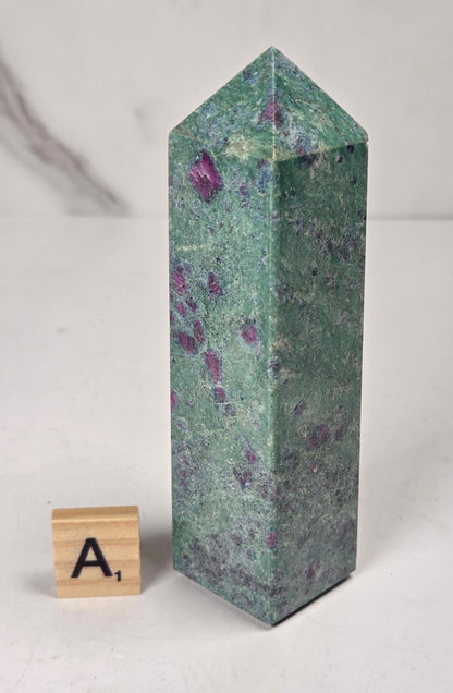Green crystal polished point with pink and purple spots next to a scrabble tile against a white background