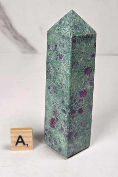 Green crystal polished point with pink and purple spots next to a scrabble tile against a white background