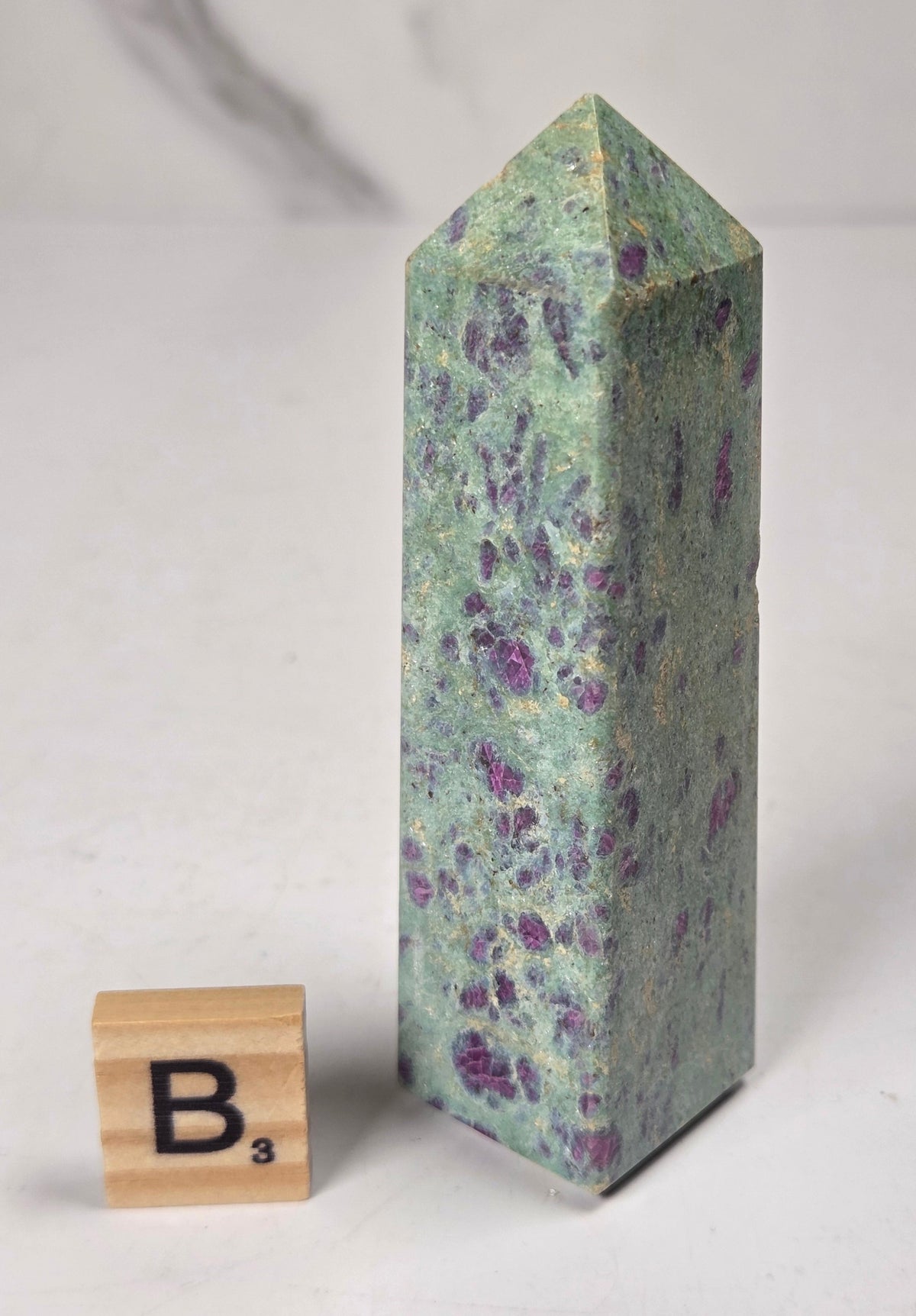 Green crystal polished point with pink and purple spots next to a scrabble tile against a white background