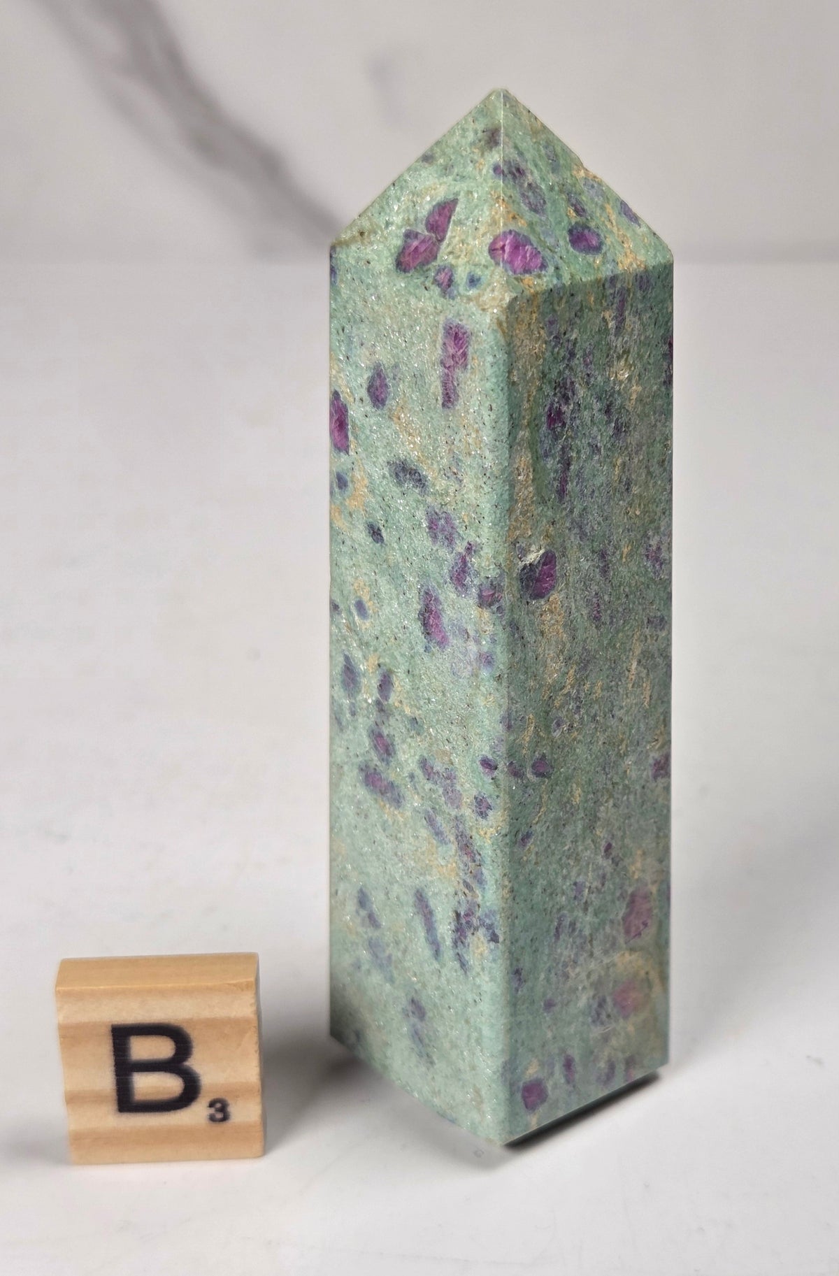 Green crystal polished point with pink and purple spots next to a scrabble tile against a white background