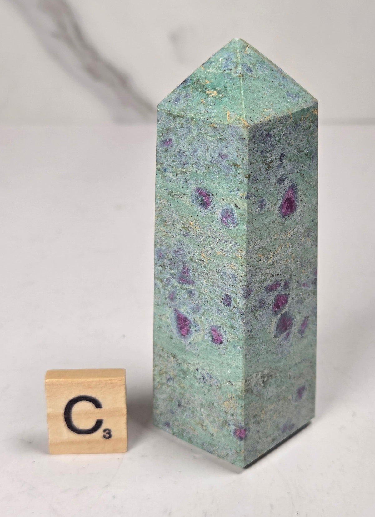 Green crystal polished point with pink and purple spots next to a scrabble tile against a white background