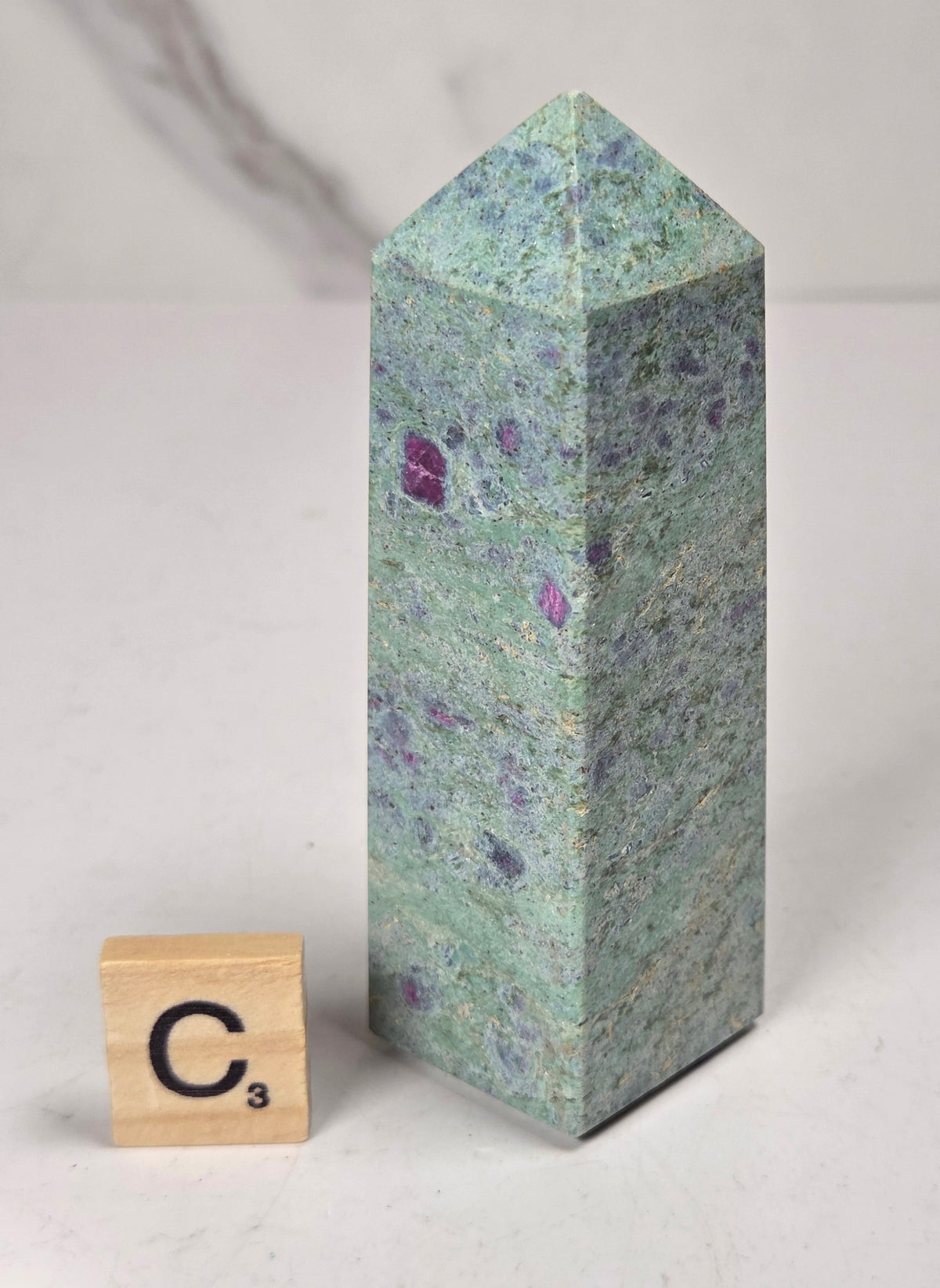 Green crystal polished point with pink and purple spots next to a scrabble tile against a white background