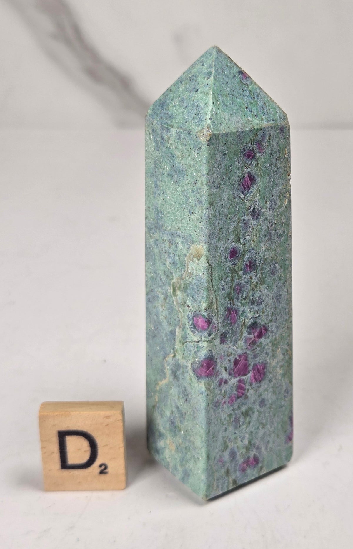 Green crystal polished point with pink and purple spots next to a scrabble tile against a white background